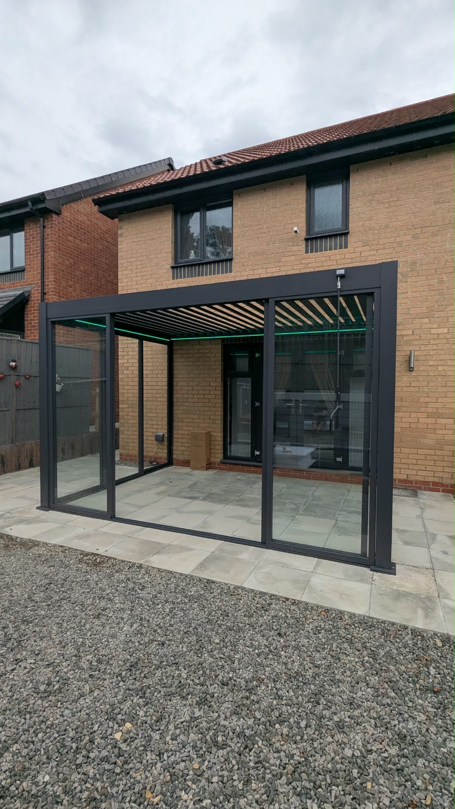 A modern glass patio canopy with a black metal frame and integrated roof louvers, installed over a paved patio area in front of a brick house.