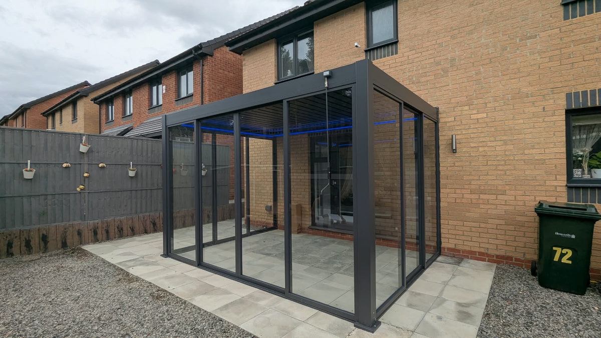 A modern glass sunroom enclosure with a dark gray metal frame installed on a paved patio in front of a brick house.